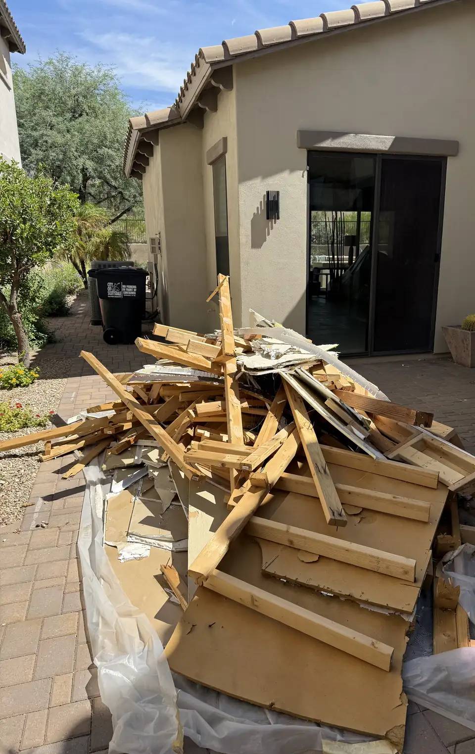 Pile of broken lumber, plywood and plastic sheeting stacked on a brick paver patio beside a house before removal