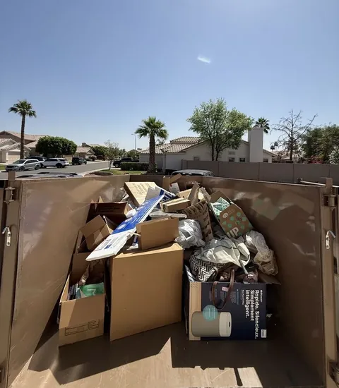 Dump trailer filled with cardboard boxes and household items, with palm trees and houses on the street behind